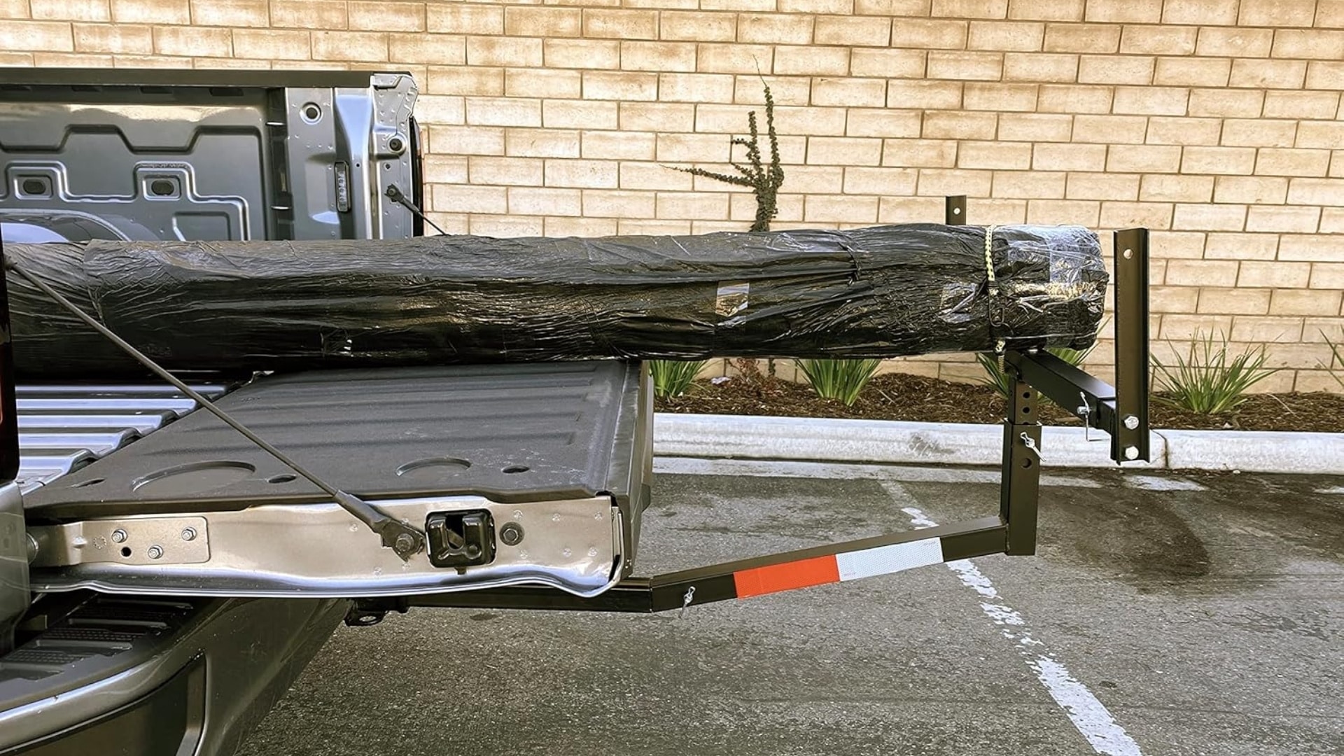 A black hitch-mounted bed extender supporting a long, wrapped cargo load in the open tailgate of a grey pickup truck.