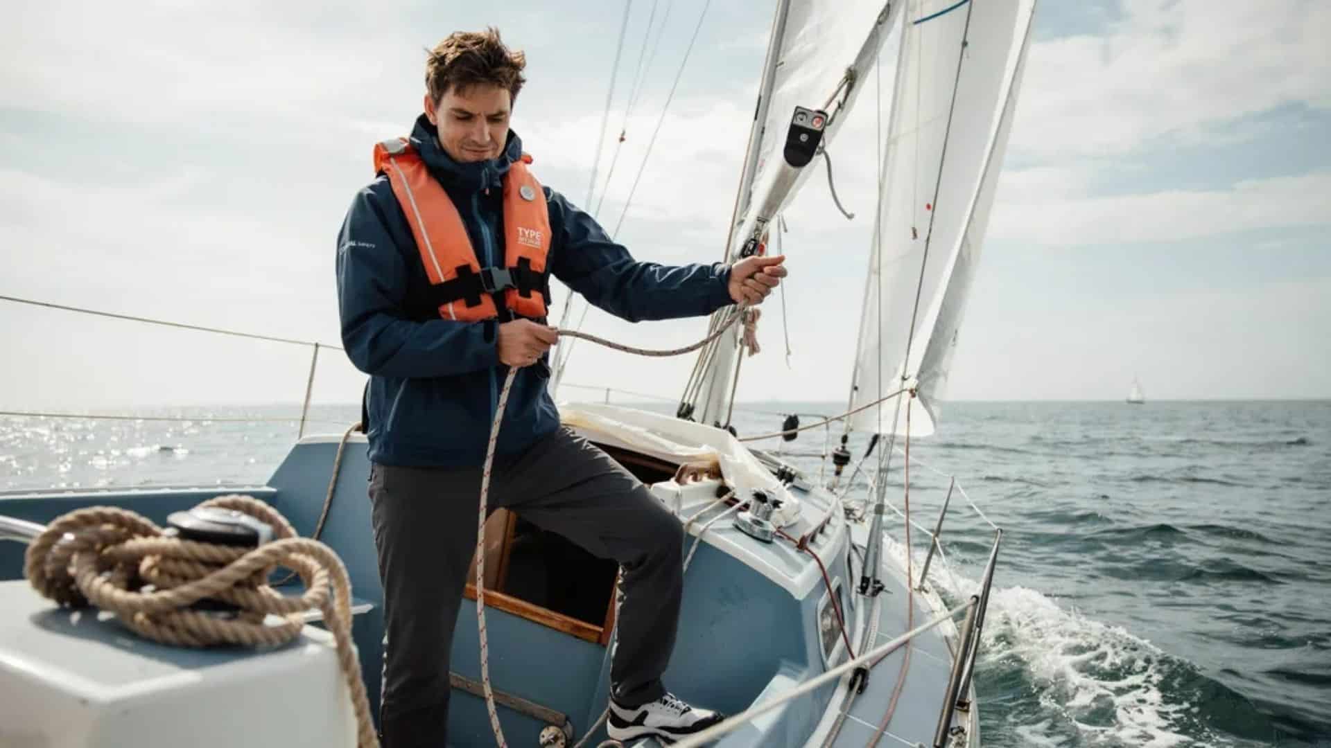 Man on sailboat wearing orange life jacket holding rope while sailing on open sea with waves.