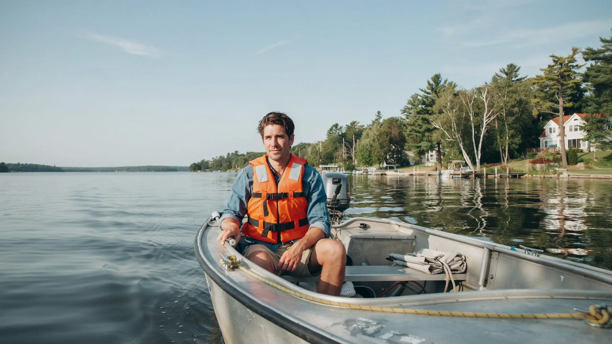 Man wearing a Type II life jacket sitting in a small boat on a calm lake near a wooded shoreline.