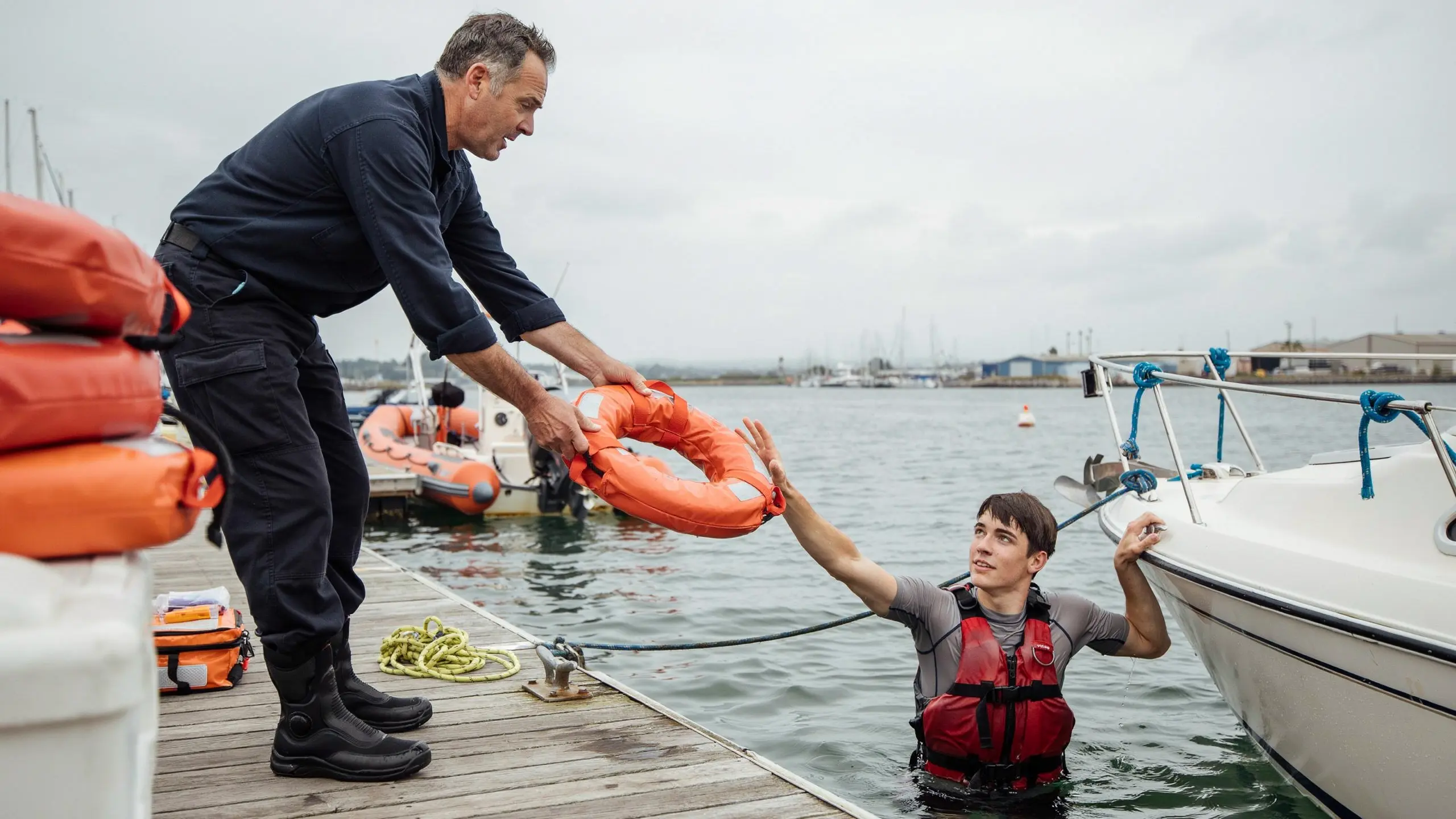 Man handing a flotation ring from a dock to a swimmer in the water beside a boat during marina rescue training.
