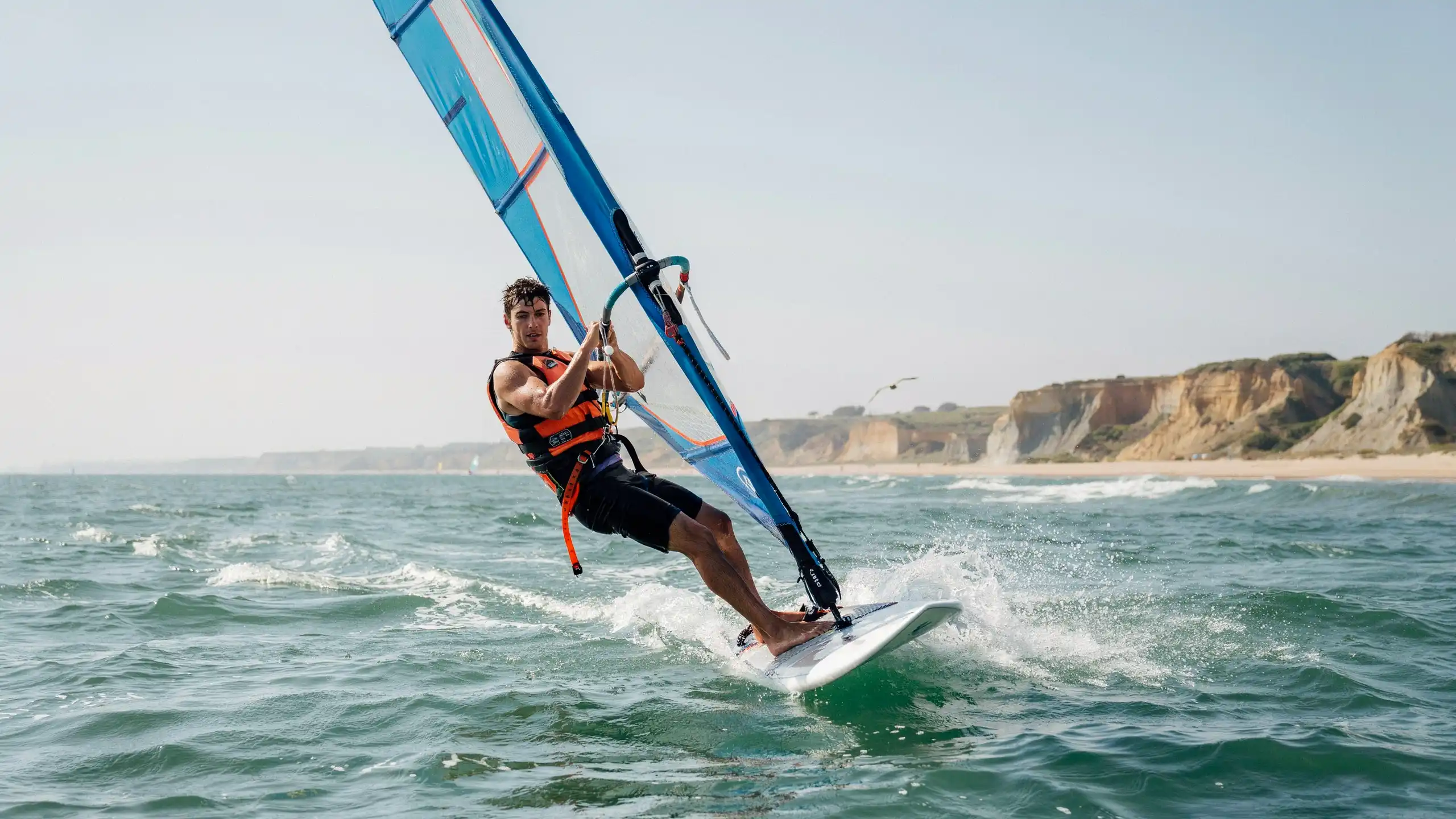 Man windsurfing in open sea wearing an orange life jacket, riding waves near a coastal cliff shoreline.
