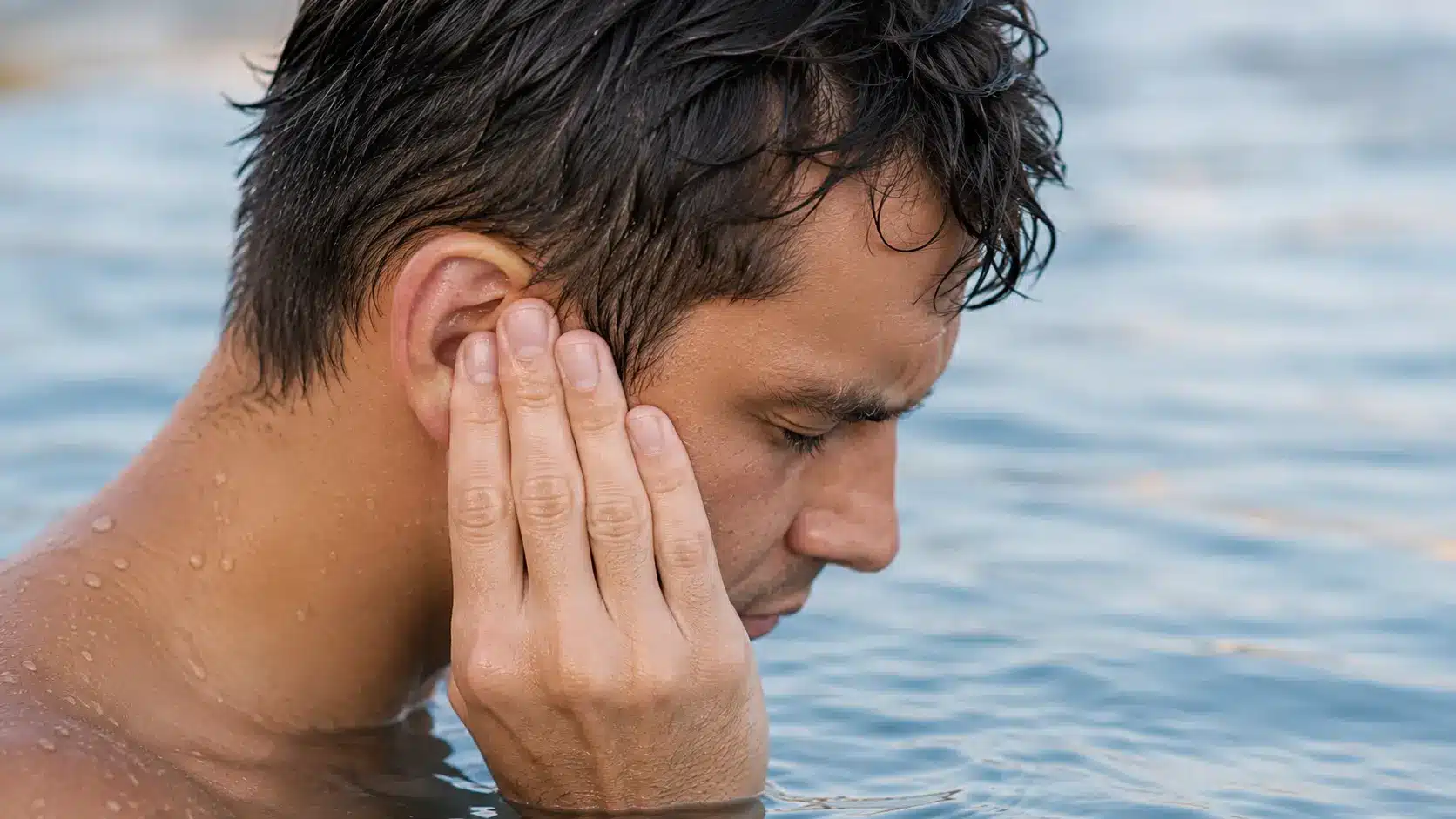 Man in water cups his ear after swimming as droplets drain, illustrating trapped water discomfort.