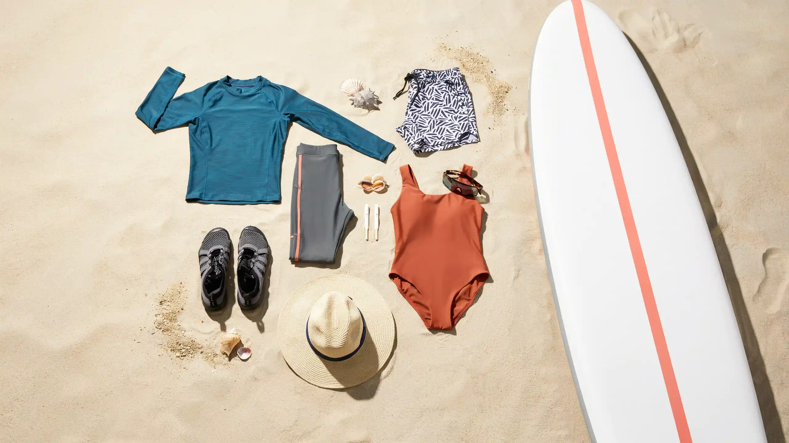 Paddle boarding gear flat lay with rash guard, leggings, swimsuit, hat, water shoes, and sunglasses arranged beside a paddle board