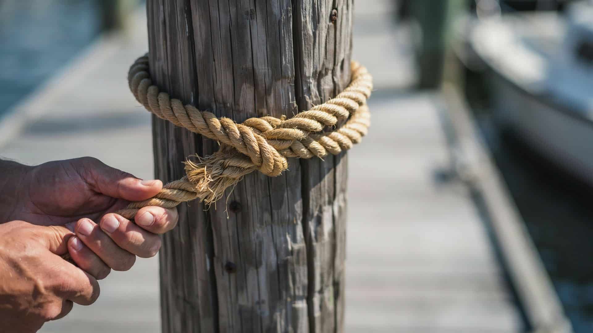 Hands tying a clove hitch knot with a thick rope around a wooden dock piling.