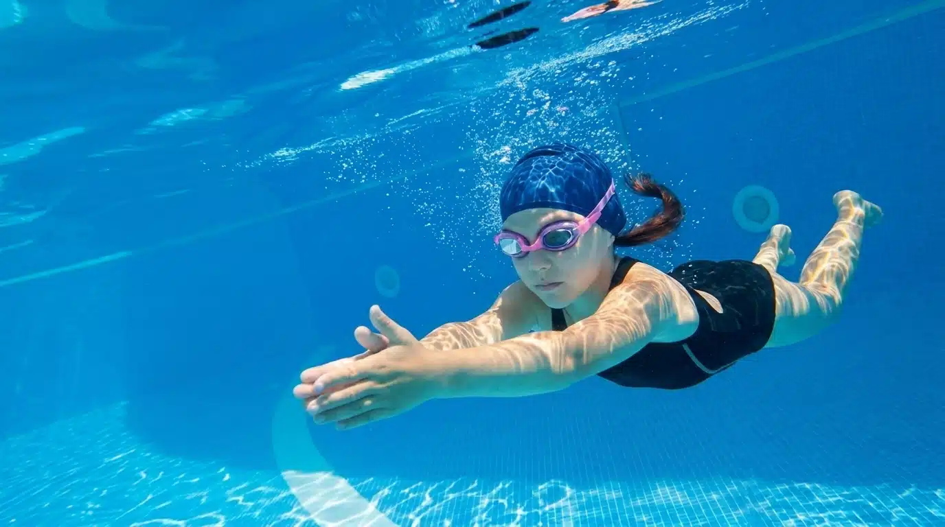 Child swimming underwater in a pool wearing goggles and a swim cap during practice.