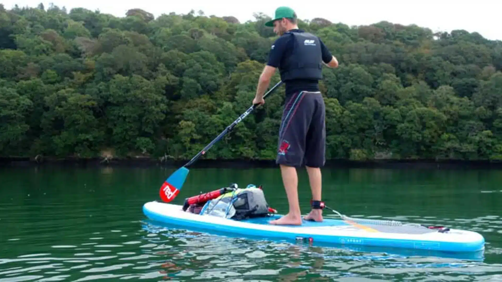 Man paddle boarding in a life vest, t-shirt, and board shorts with gear strapped to the board near a wooded shoreline
