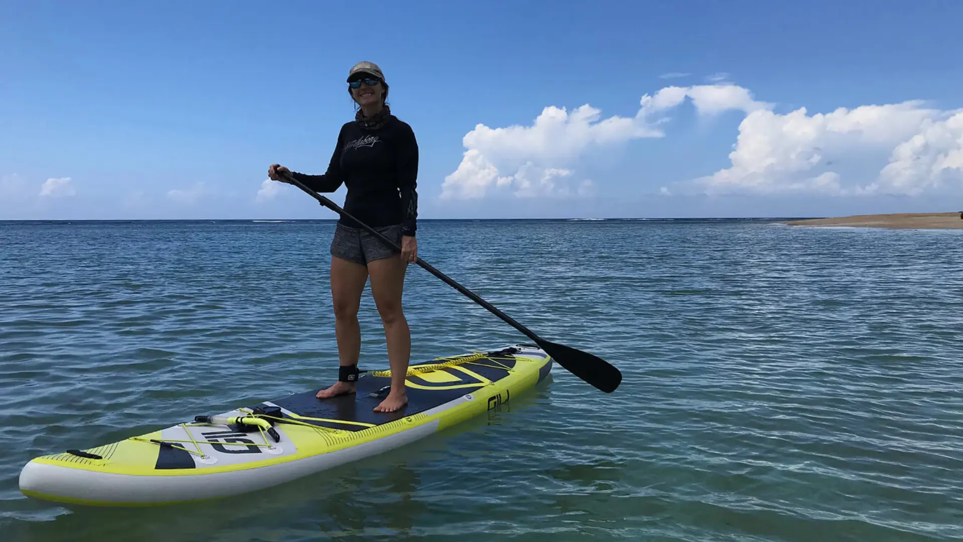 Woman paddle boarding in a long-sleeve rash guard, patterned shorts, cap, and sunglasses on a calm ocean