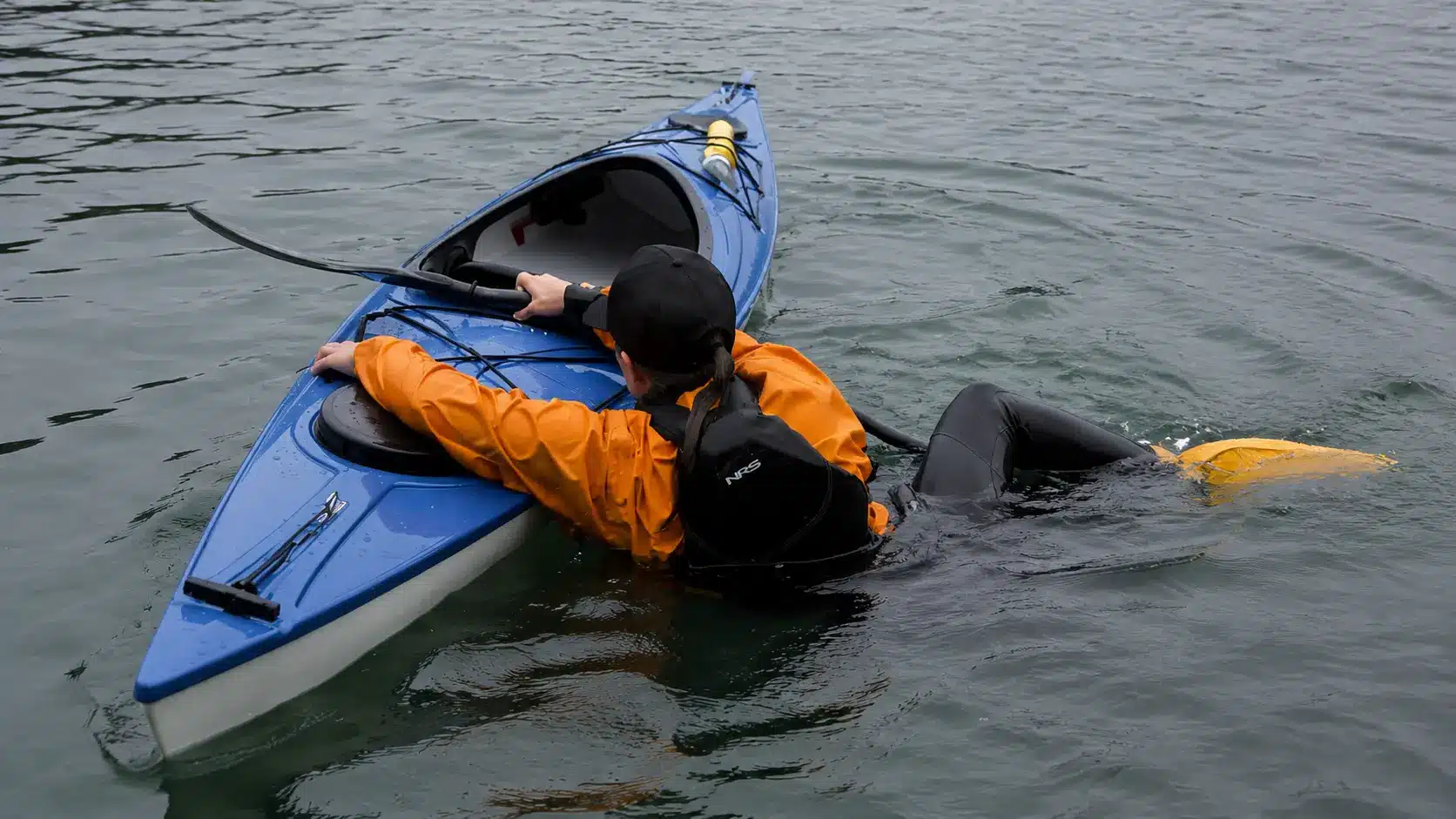 Kayaker struggling to re-enter a capsized blue kayak in calm water during a paddle float rescue attempt