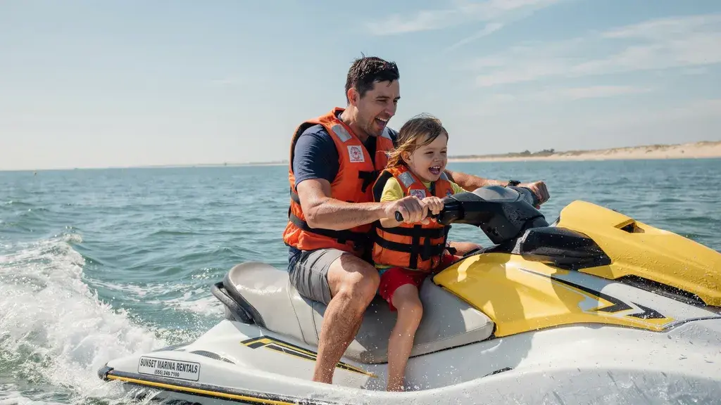 Parent and child wearing life jackets ride a yellow jet ski together on open water during a sunny ocean adventure.