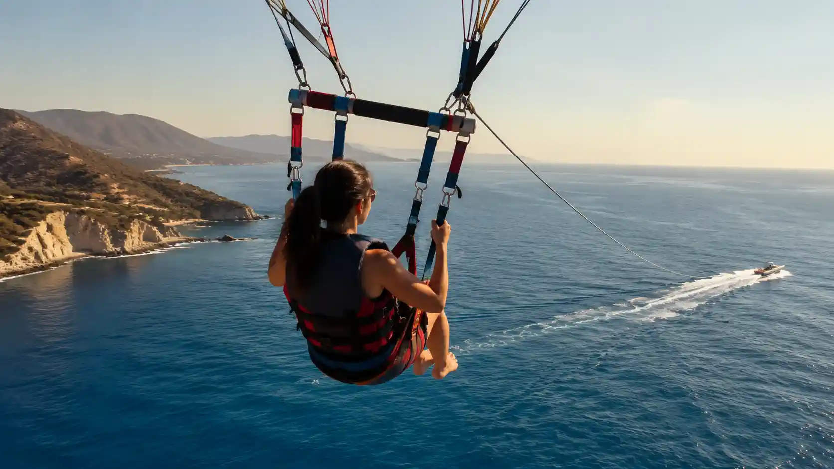 Wide-angle realistic aerial view of a parasailing boat towing a single rider over calm turquoise ocean with distant cliffs under clear sky.