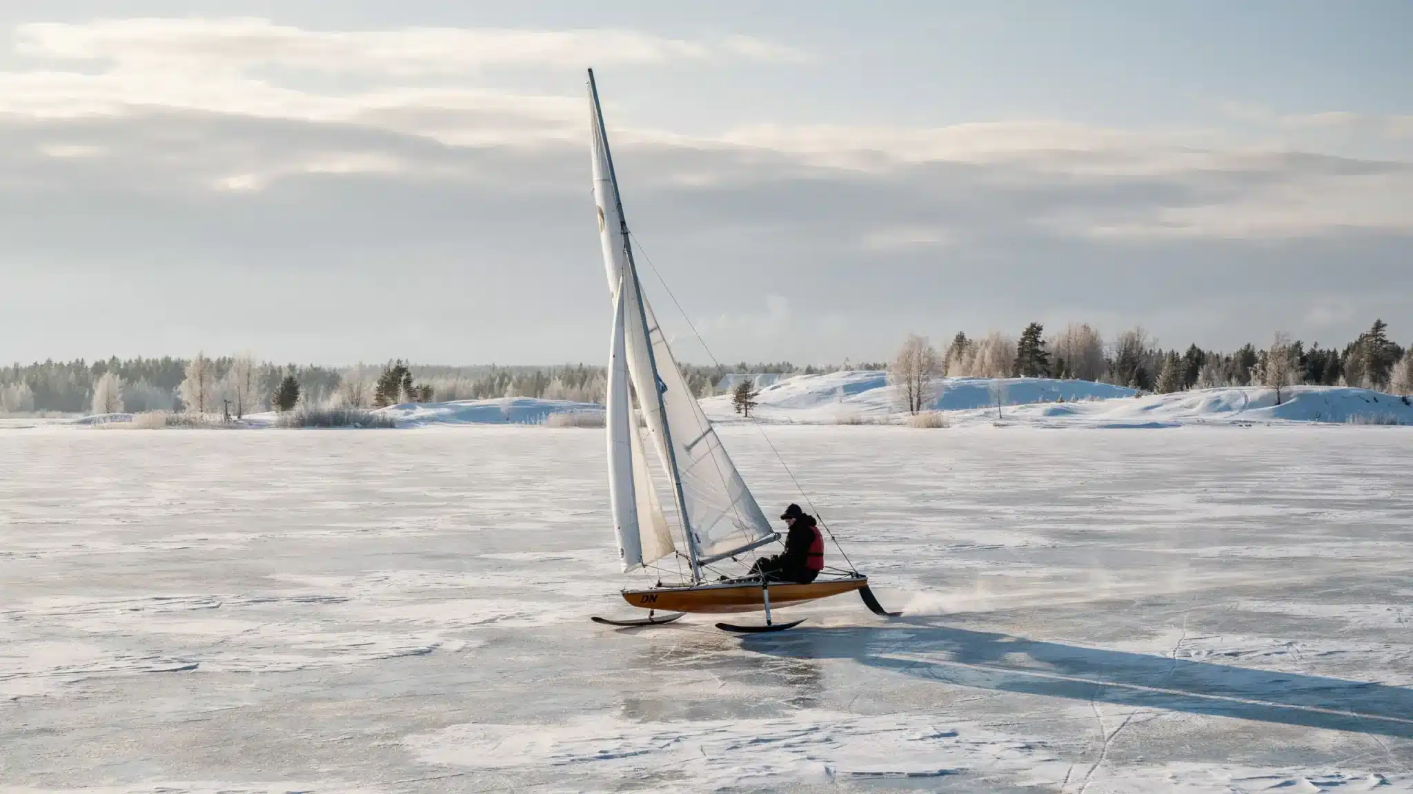 Compact DN iceboat racing across a frozen lake with sail raised and runner blades visible