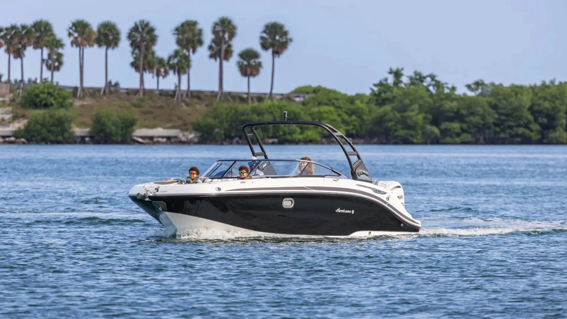 Black and white deck boat cruising on calm water with wake tower and scenic shoreline surroundings