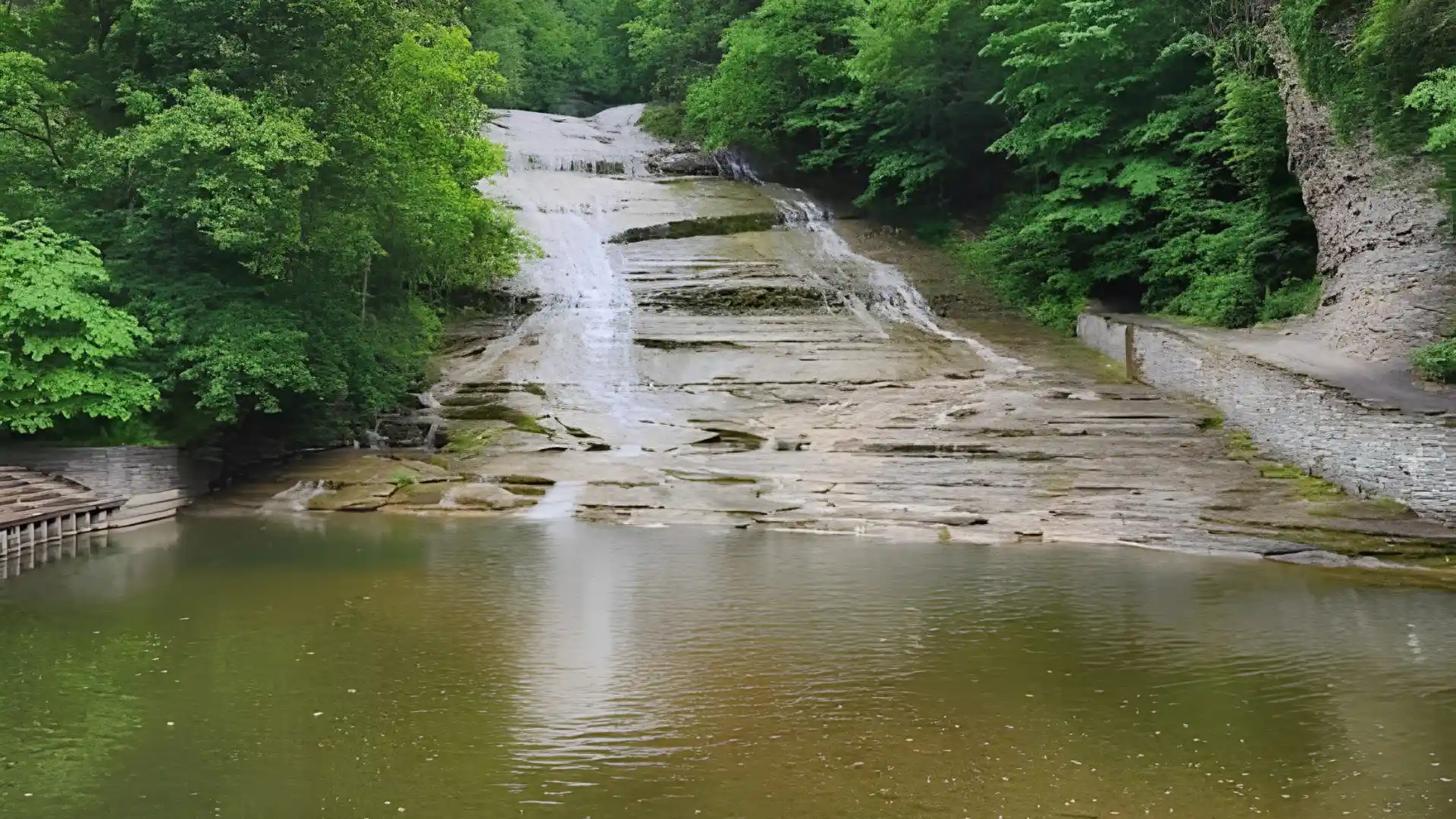 Broad cascade spreads across layered stone ledges before reaching a quiet pool surrounded by lush woodland and walking paths