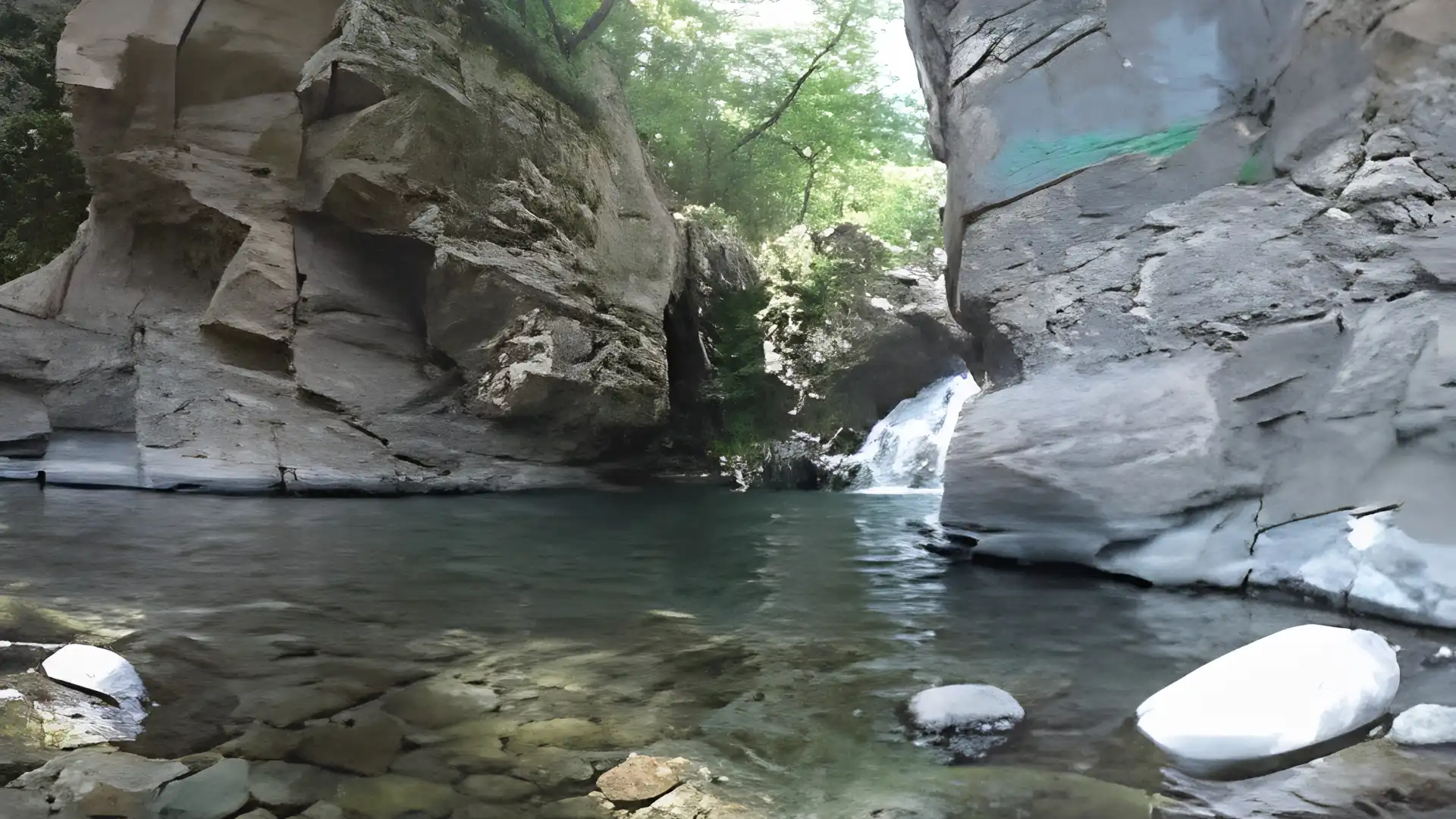Clear rock pool between towering gray canyon walls with a small waterfall flowing beneath a leafy forest canopy