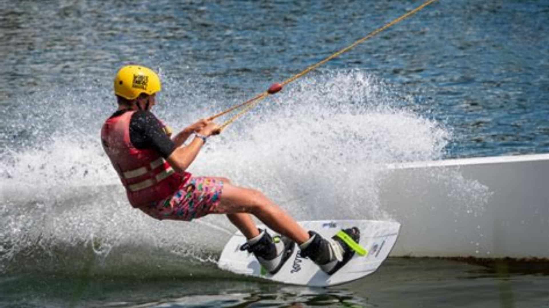 Wakeboarder riding behind a boat showing proper board size, balance, and control on a calm lake with water spray