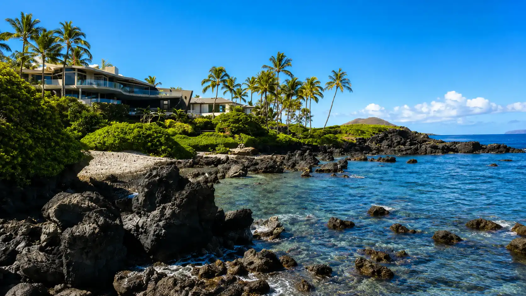 Rocky tropical coastline with palm trees, clear blue ocean, lush greenery, and beachfront resort under a sunny sky