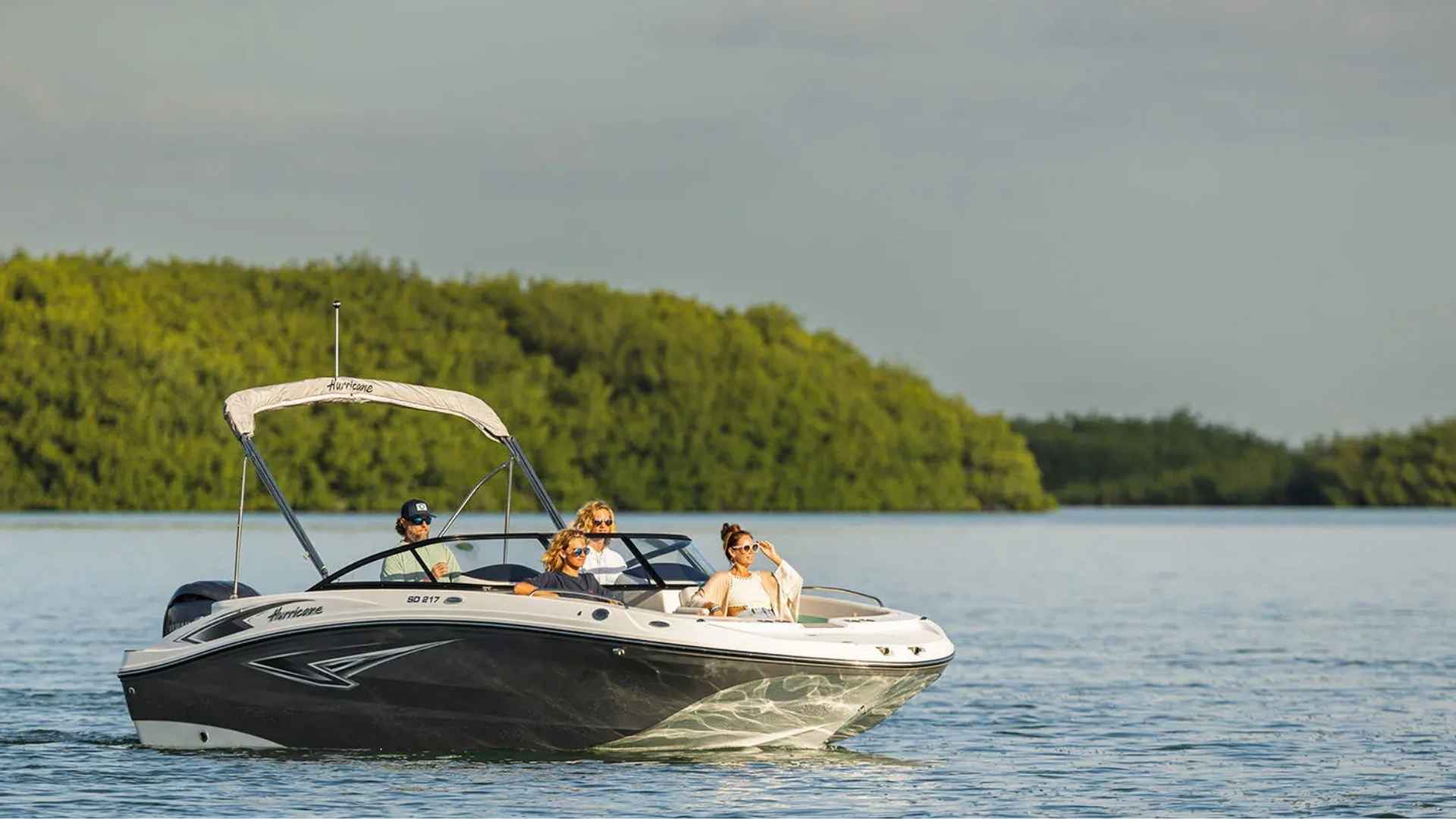 Hurricane SunDeck 217 OB black and white deck boat cruising on calm water with passengers and green shoreline background