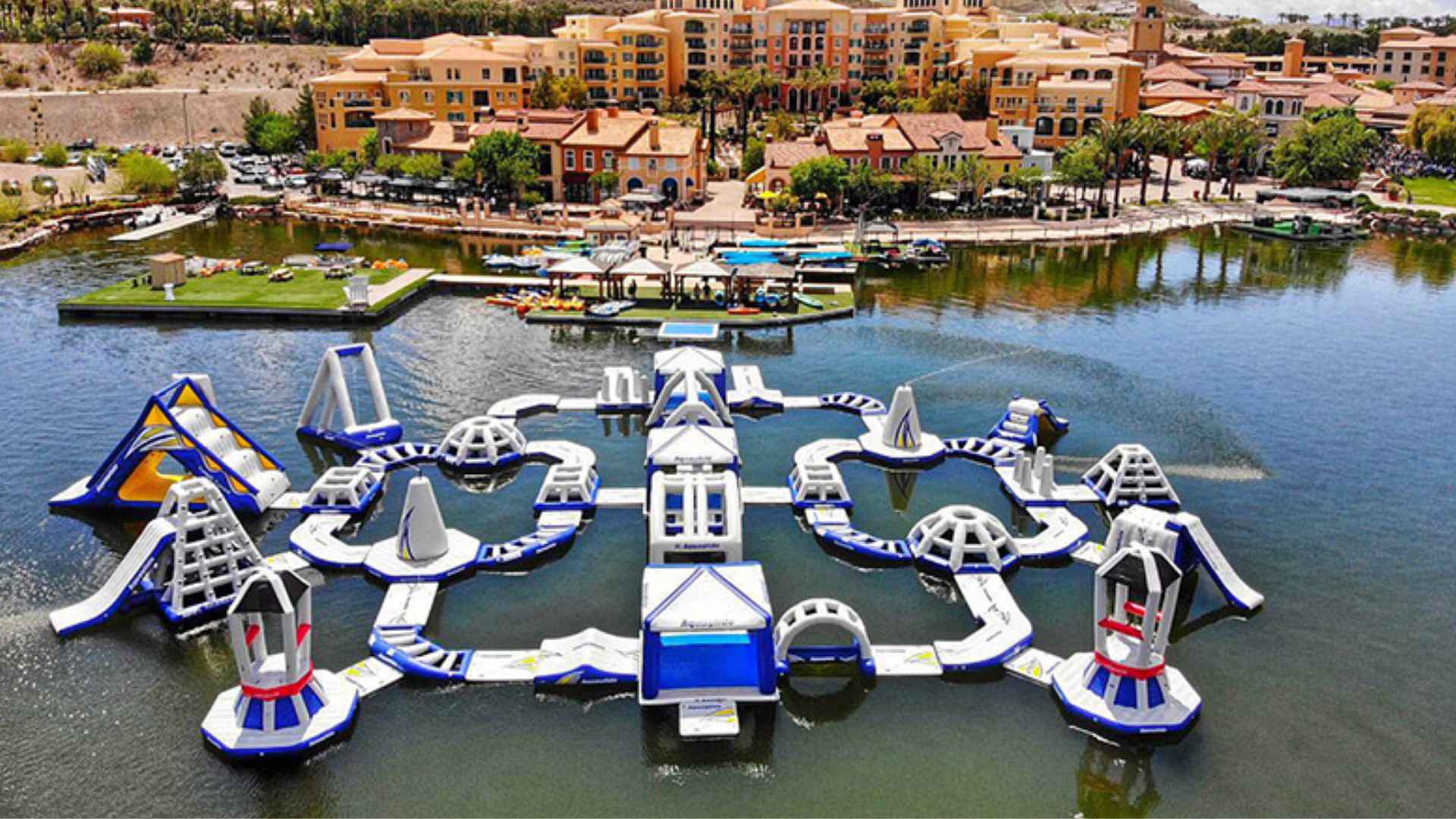 Inflatable aqua park floating on Lake Las Vegas with colorful resort buildings, docks, and calm water in the background
