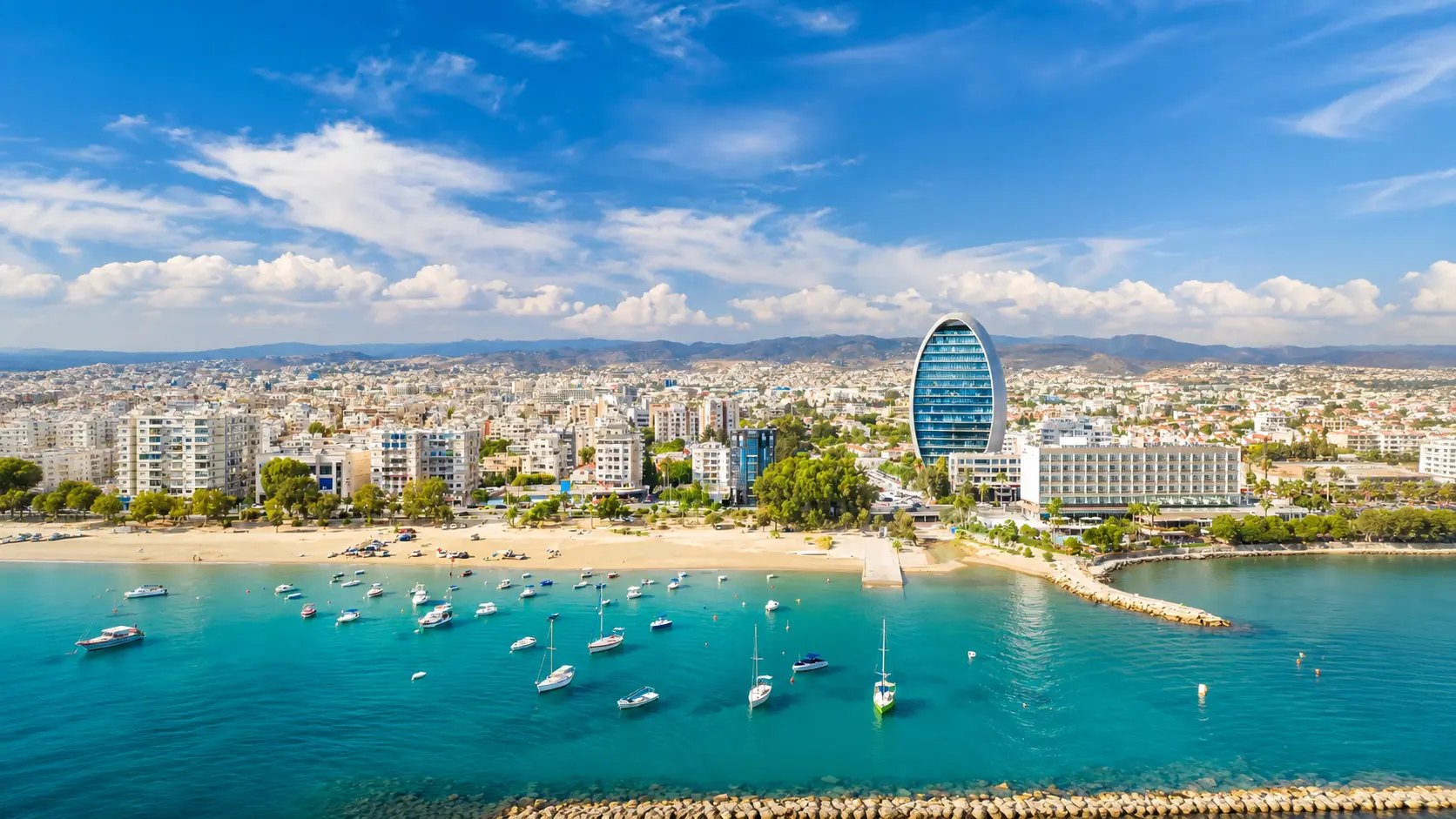 Aerial view of Larnaca with turquoise bay boats sandy beach waterfront buildings and mountains under blue sky
