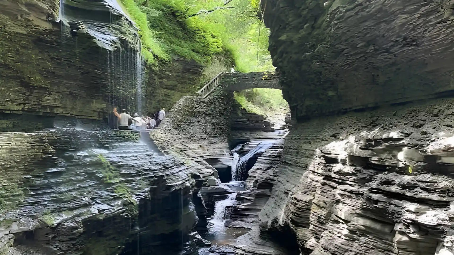 Layered stone gorge with narrow waterfalls, a rustic footbridge, and visitors walking beside the shaded stream