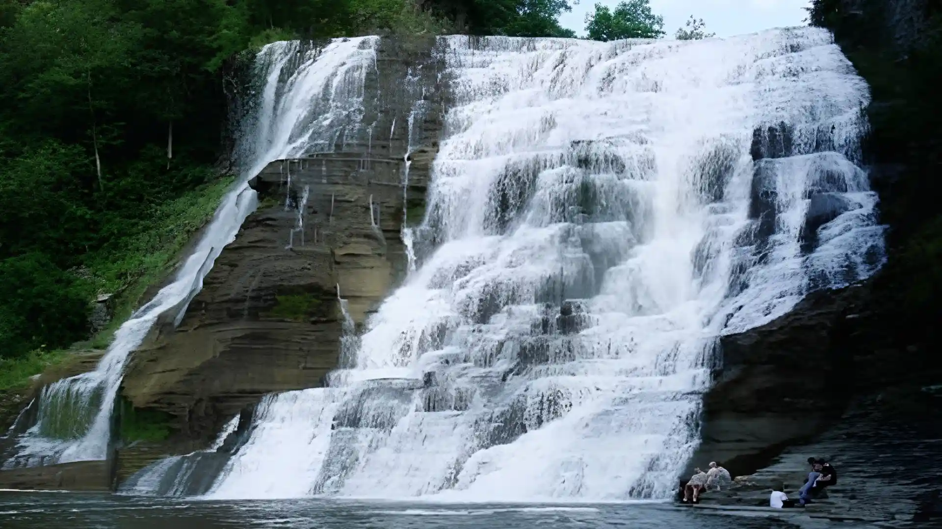 Massive cascade spreads across stepped stone formations with white water pouring into a calm forest pool below