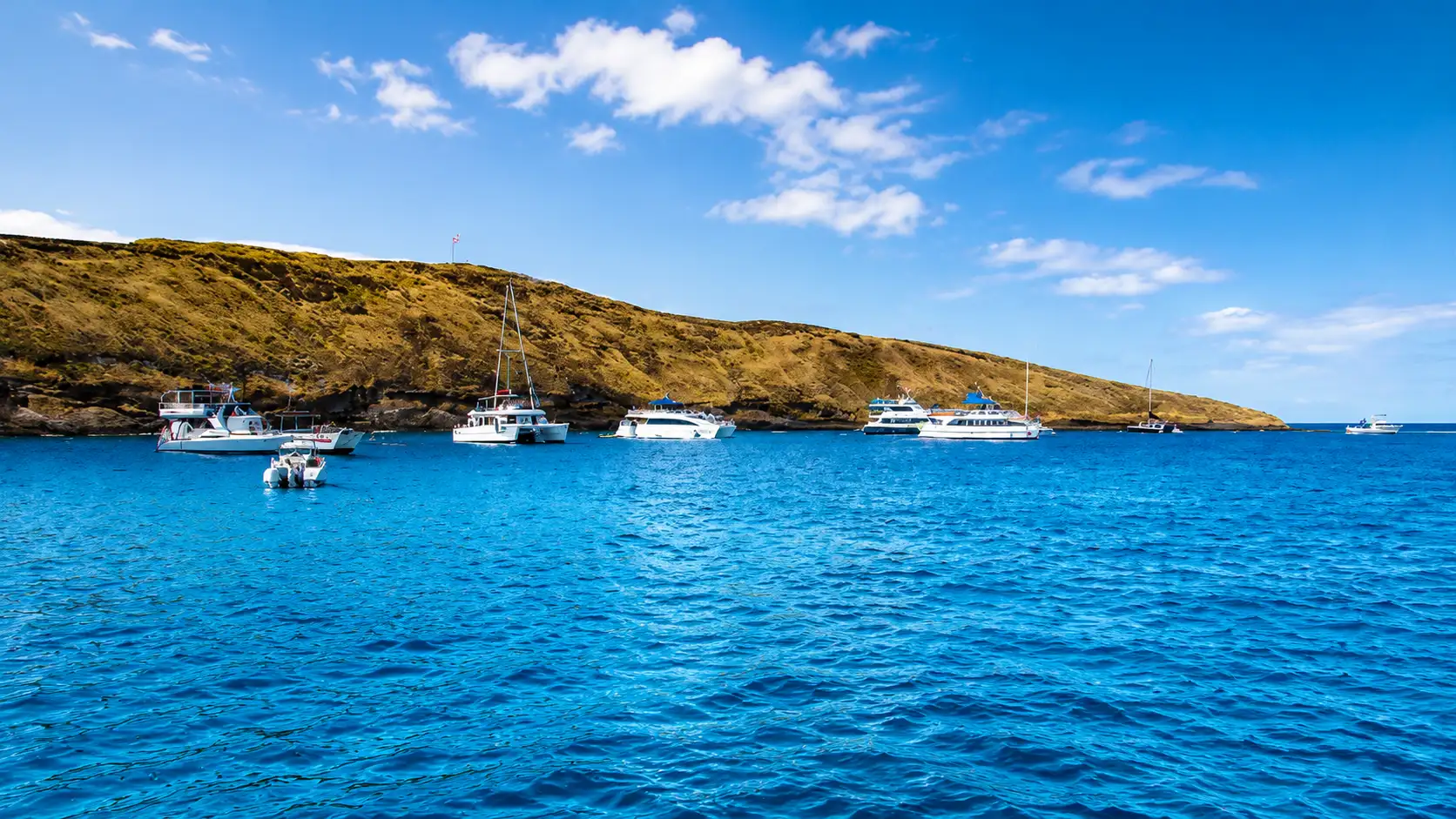 Boats anchored in a blue coastal bay with calm ocean waters, rocky hills, and a bright sky over the shoreline