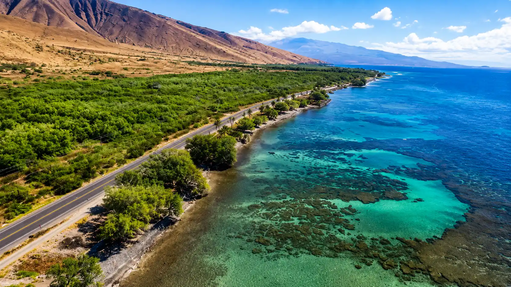Aerial coastal view showing turquoise ocean waters, rocky shoreline, green vegetation, mountain slopes, and a scenic road route