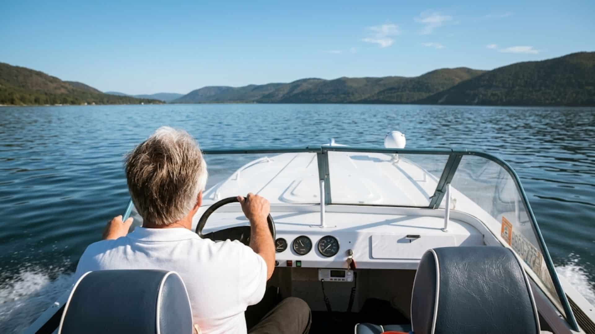 Person steering a motorboat across a calm lake with mountains and clear blue sky in the distance.