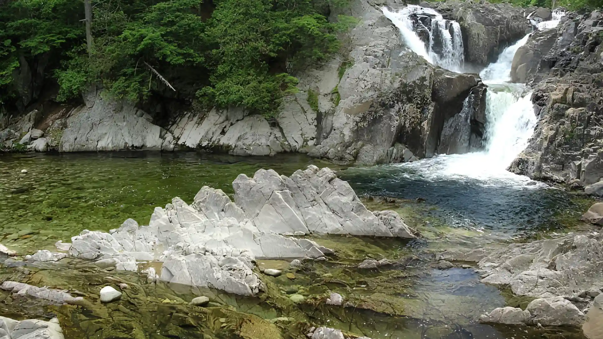 Scenic multi level waterfall tumbles over dark boulders into a transparent pool framed by sharp rocks and green trees