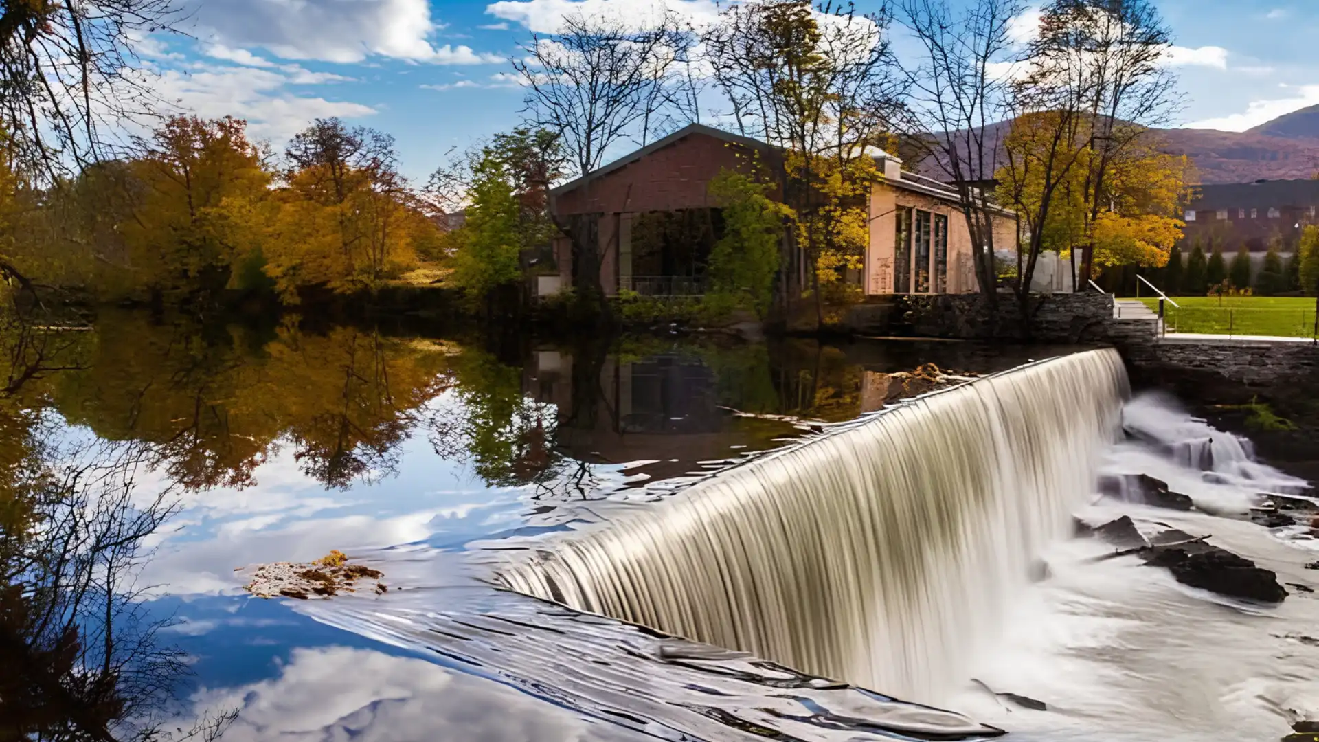 Scenic riverside waterfall flowing past a historic brick building framed by golden autumn trees and distant mountain slopes