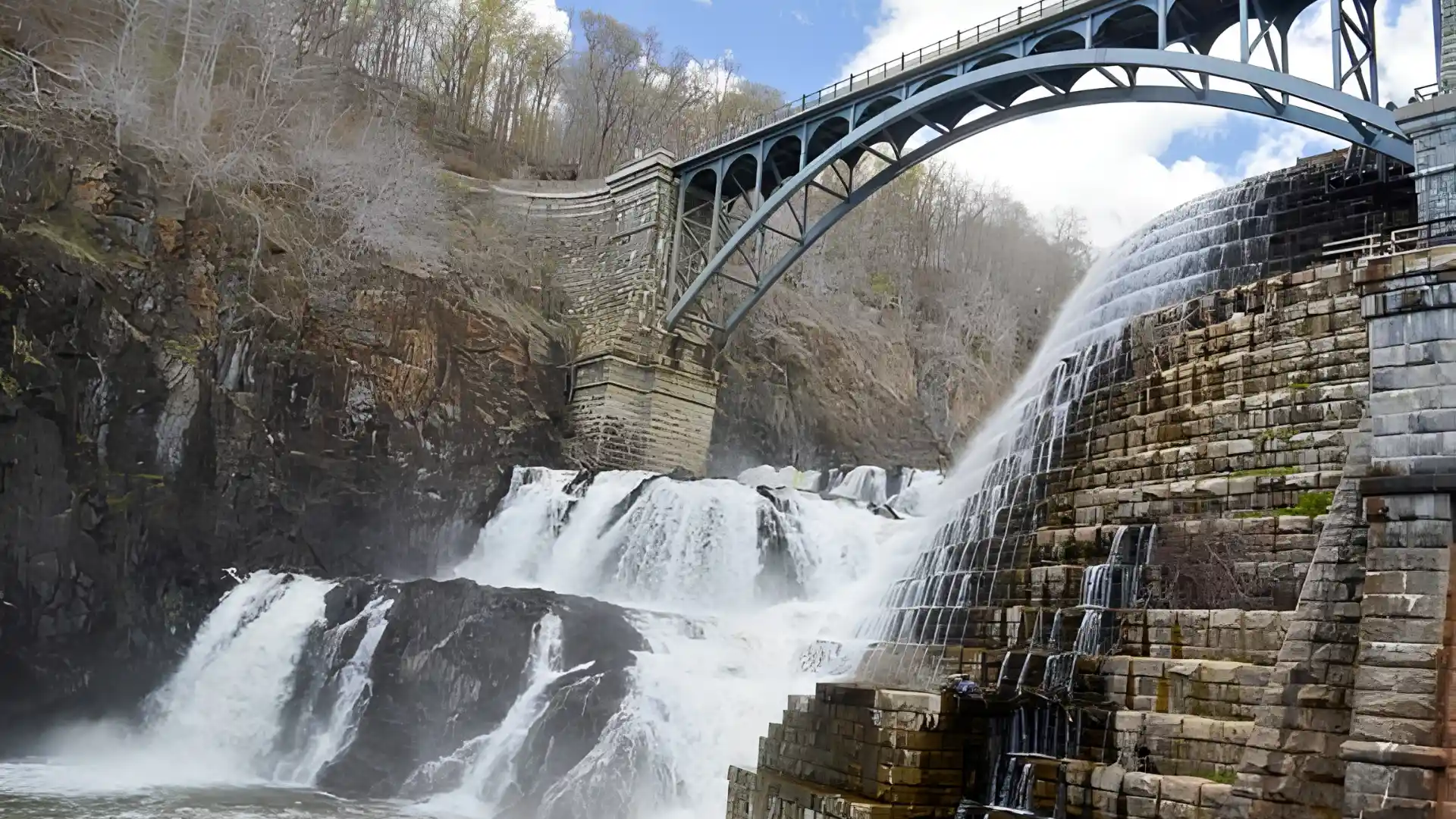 Steel arch bridge spans a powerful tiered waterfall flowing over a historic stone dam within a steep wooded gorge