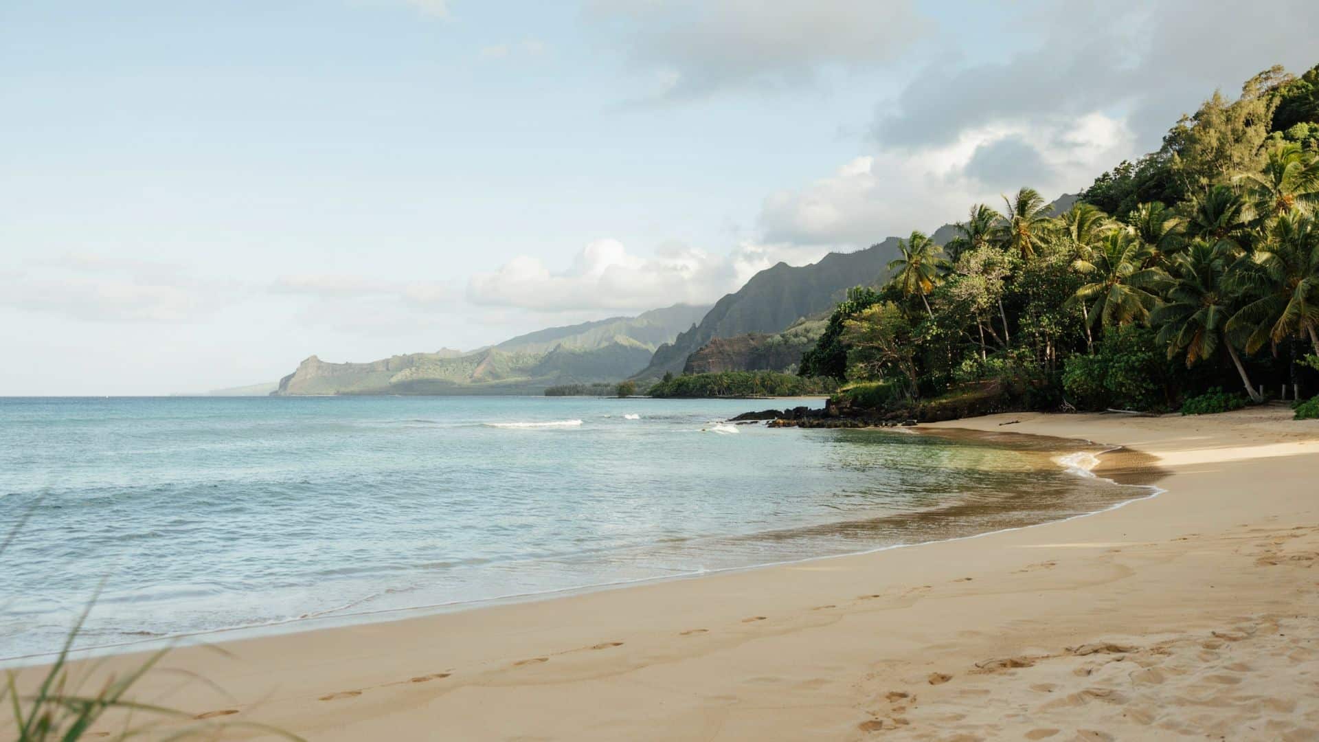 Tunnels Beach featuring a quiet sandy shoreline, blue ocean waves, tropical forest, and rugged mountain scenery