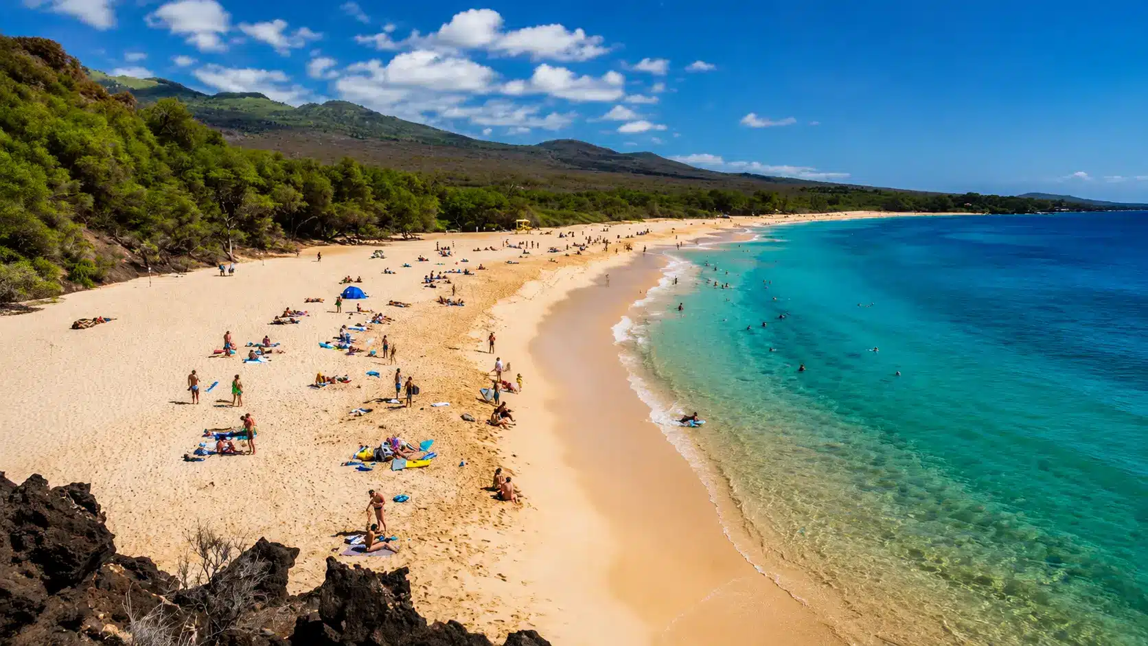 Aerial view of a tropical beach with golden sand, turquoise ocean, swimmers, green hills, and a clear blue sky