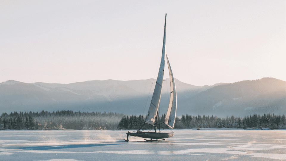 Iceboat sailing across a frozen lake with mountains and snowy forest in the background