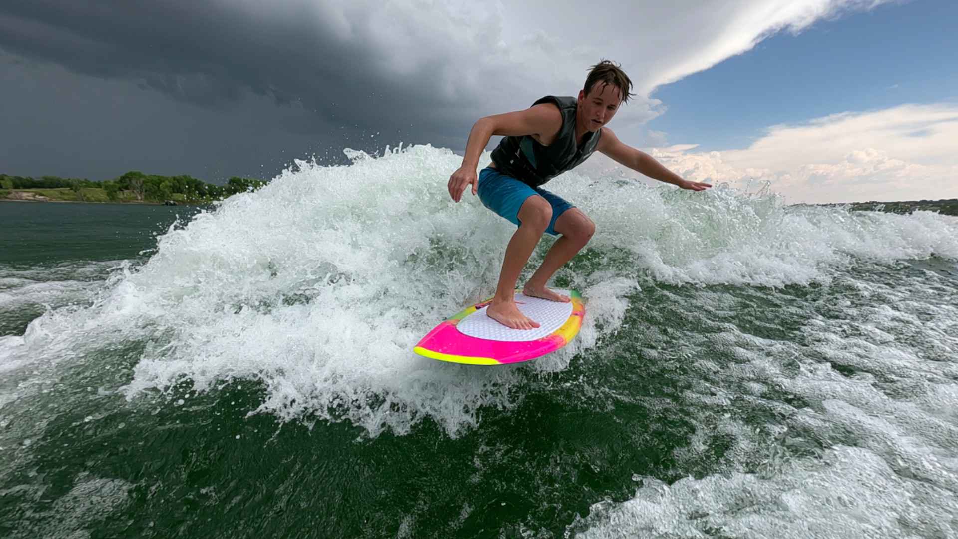 Man wakesurfing behind a boat on a colorful board, carving through white water beneath dark storm clouds and blue sky