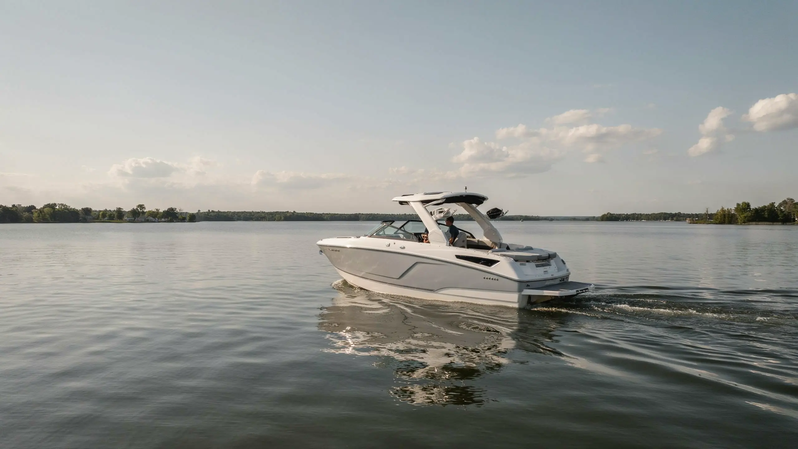White wakeboarding boat cruising across calm lake water with a spacious deck and clear blue sky in background