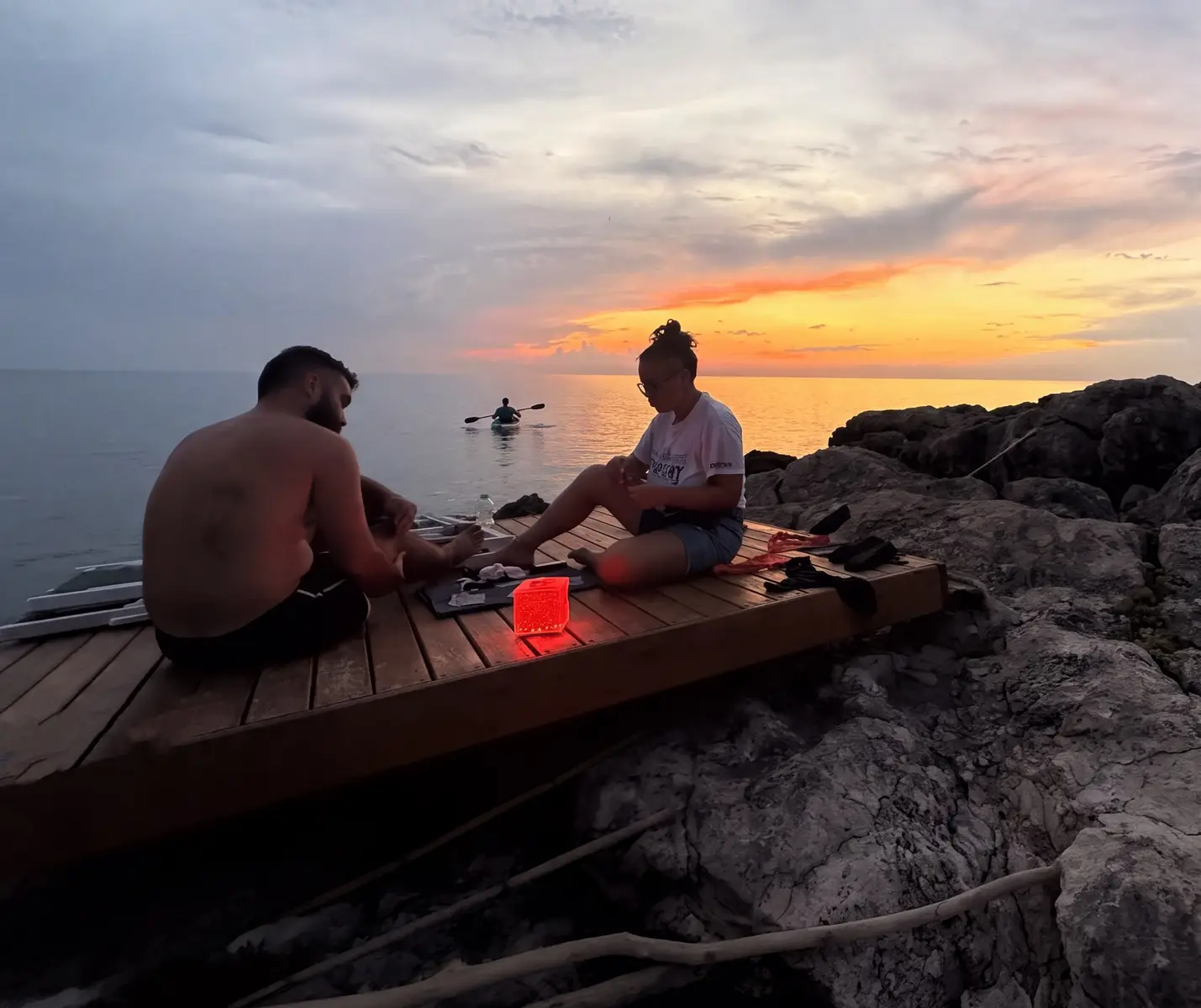 Couple sitting on wooden deck by the sea at sunset with kayaker in background