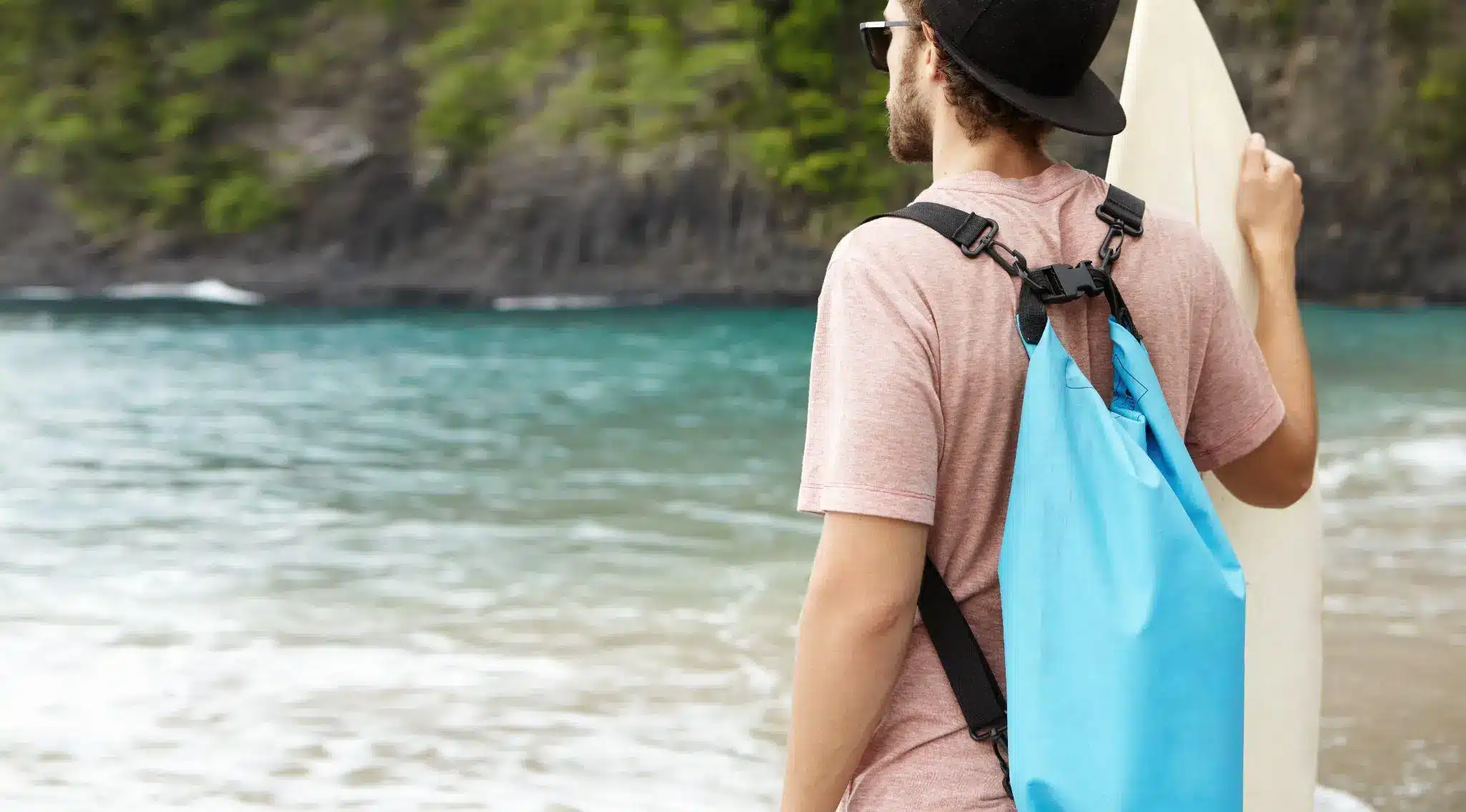 Man in hat holding surfboard by the ocean with cliffs and trees in the background