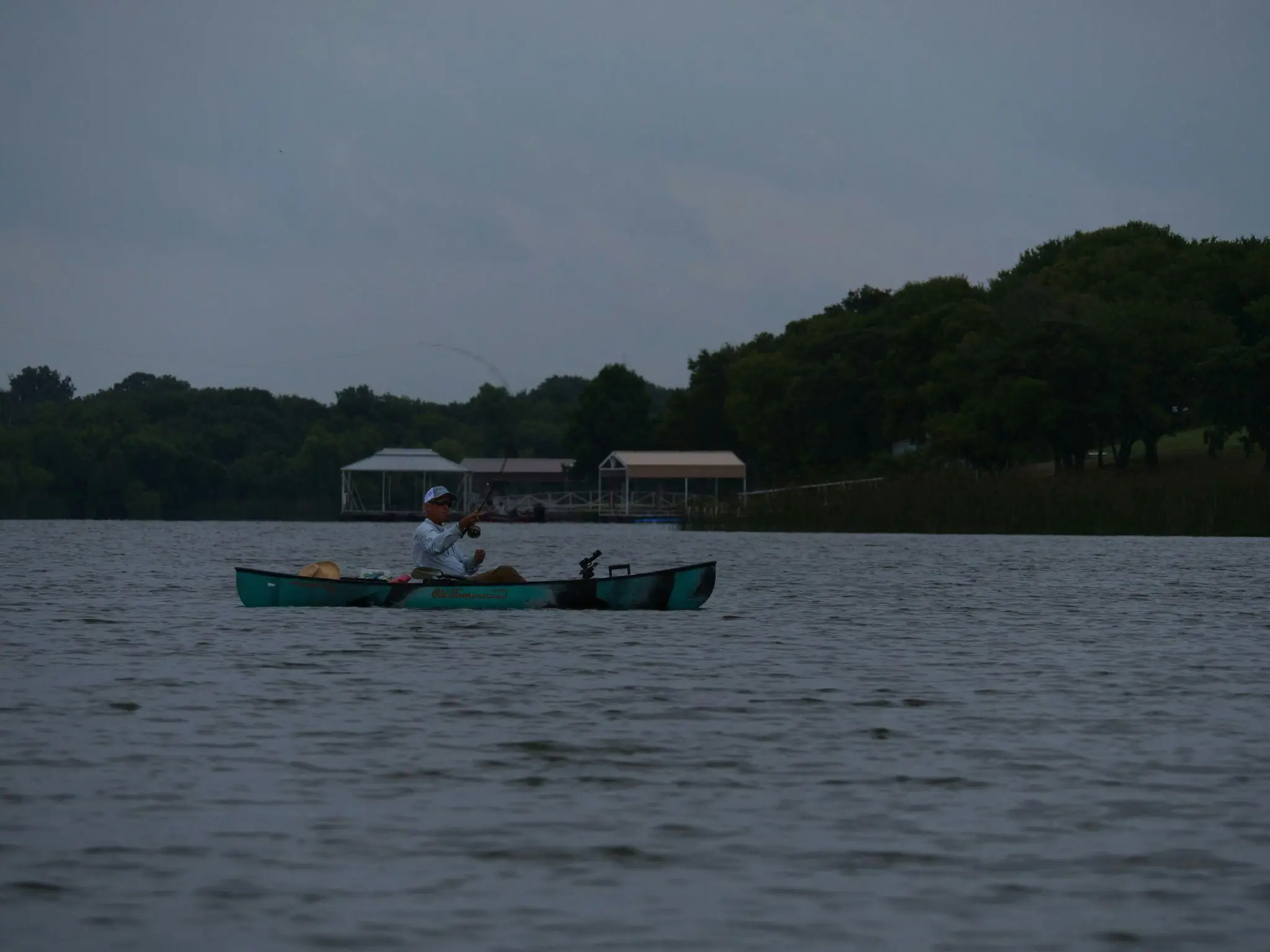 Person fishing from turquoise kayak on calm lake with tree-lined shore in background