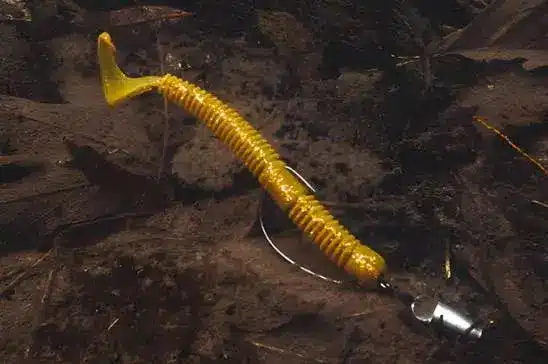 A rigged yellow paddle-tail texas rig with a hook and silver bullet weight rests on dark soil.