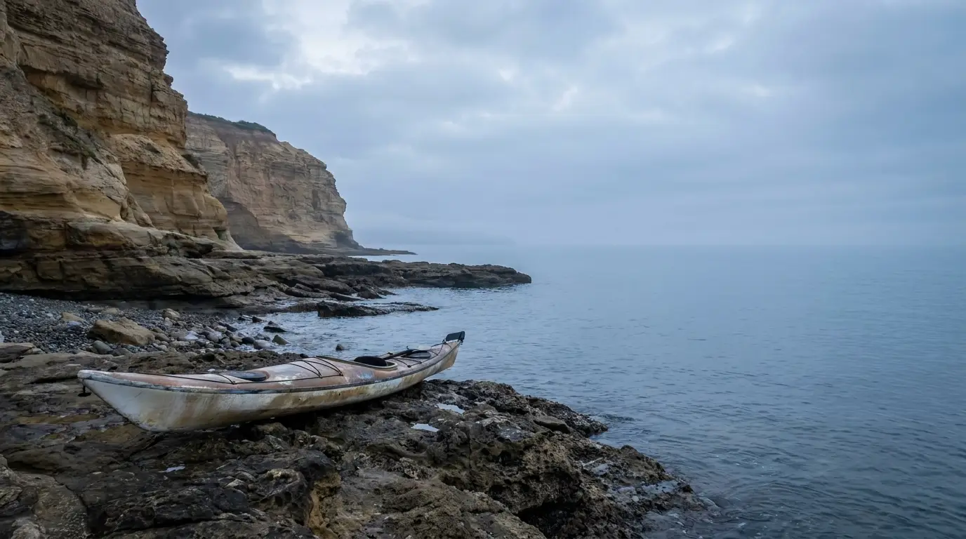 Weathered kayak on rocky shore near towering cliffs under cloudy sky