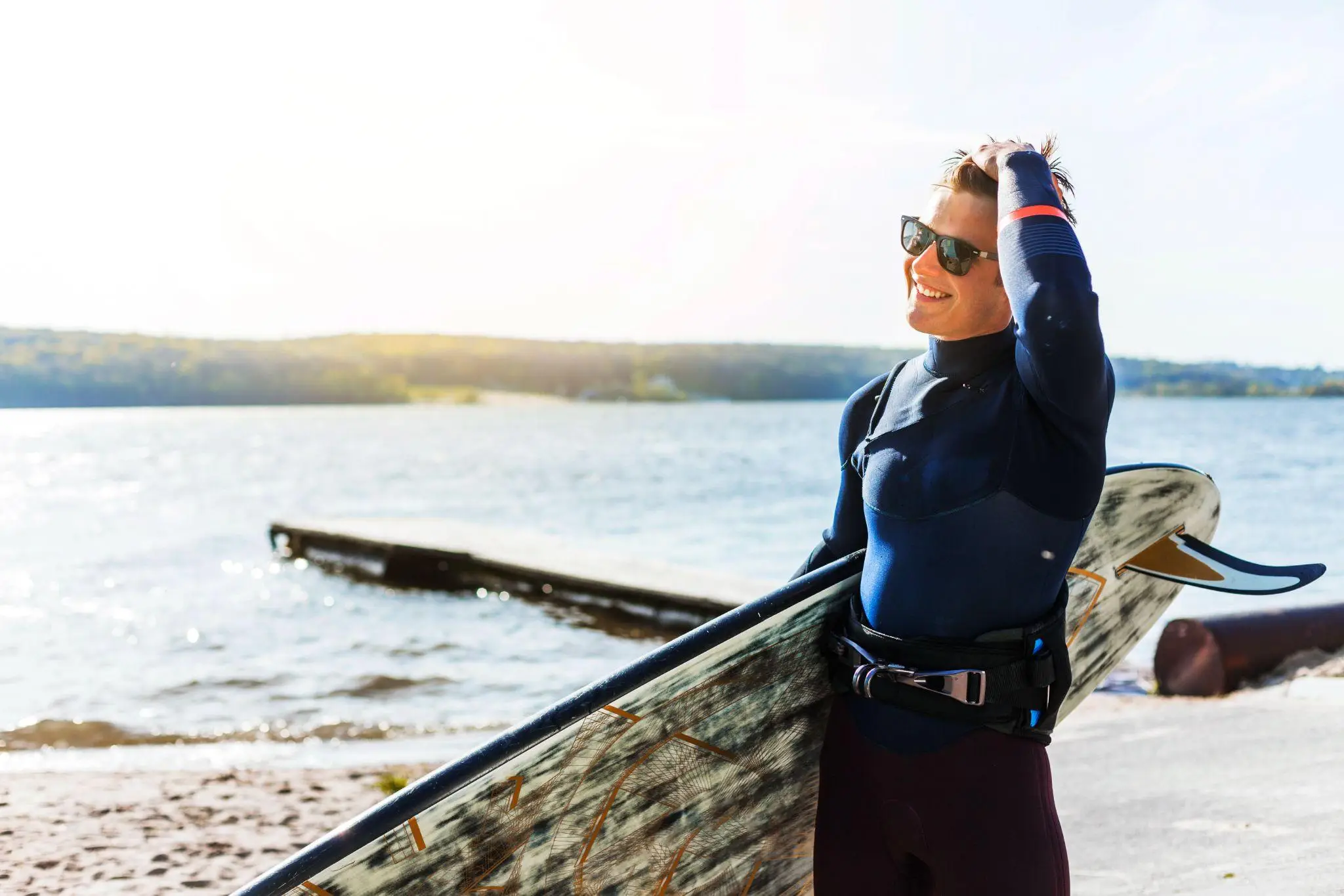 Surfer in wetsuit holding surfboard on sunny lakeshore with smiles and sunglasses