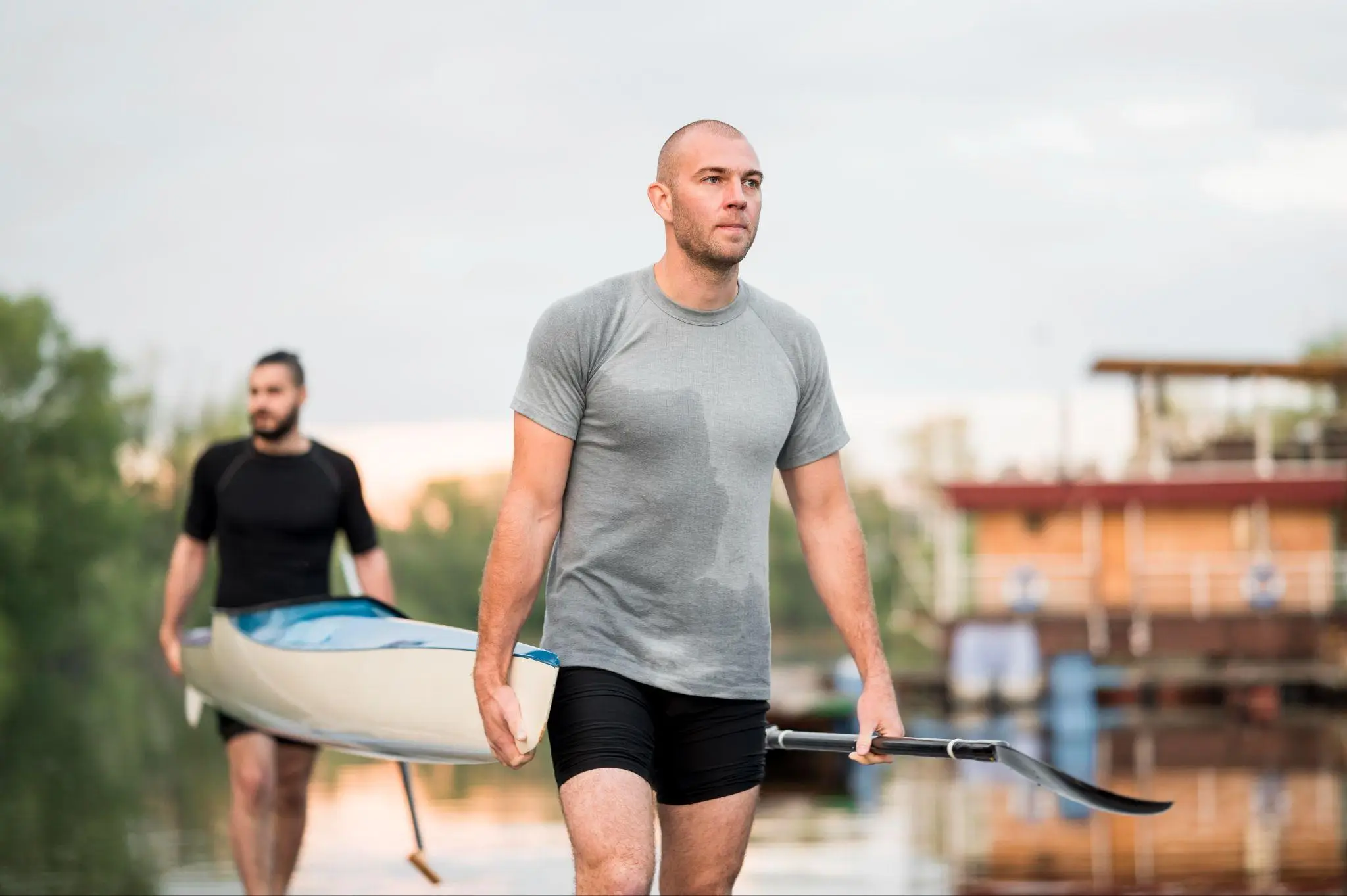 Two men carrying kayak near river with blurred trees and buildings in background