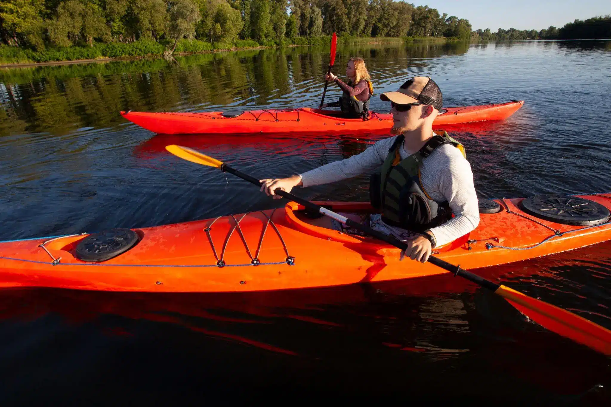 Two people kayaking in bright orange kayaks on a calm lake with lush green trees