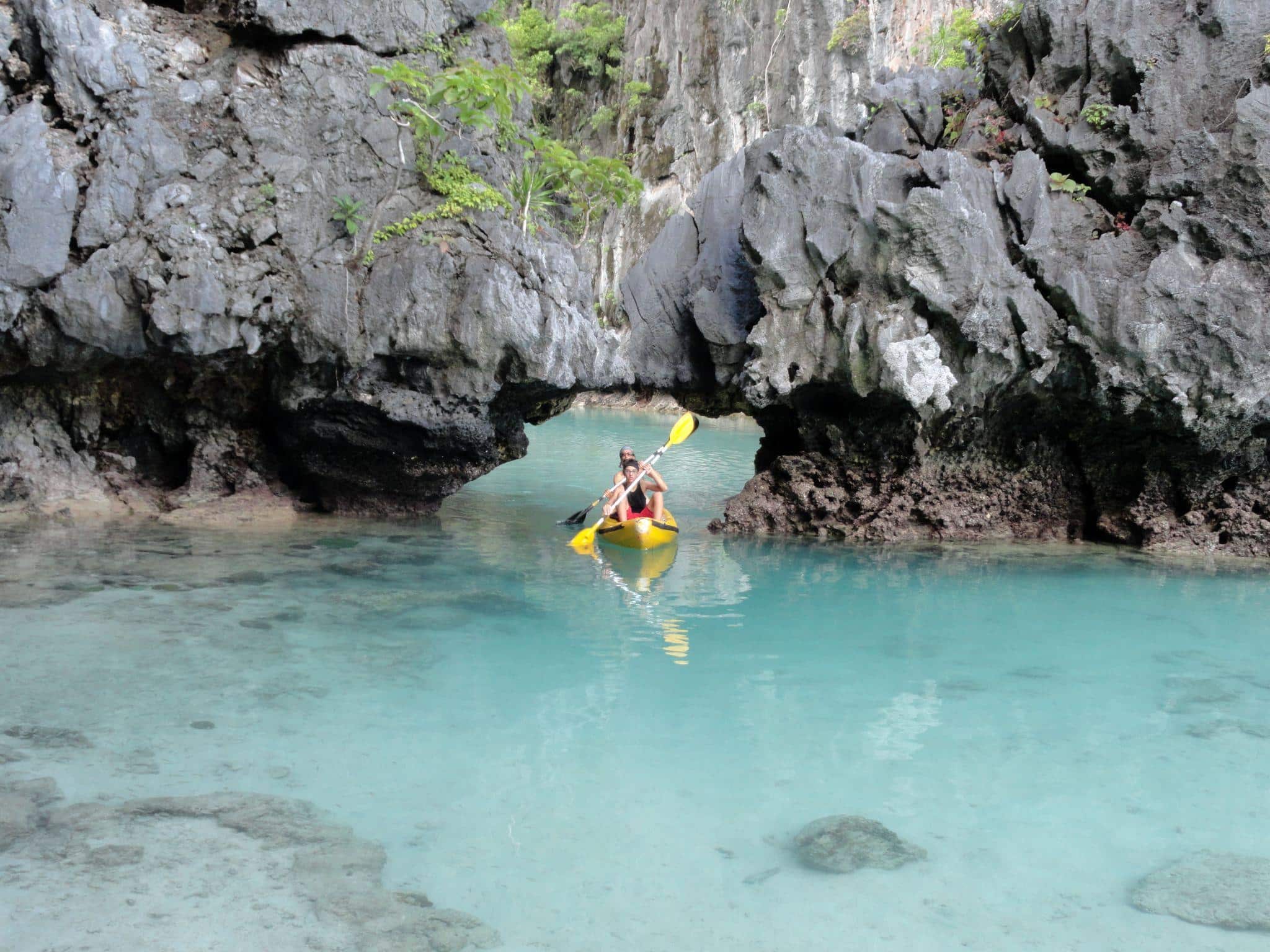 Kayaker paddling through rock archway in clear turquoise water surrounded by cliffs