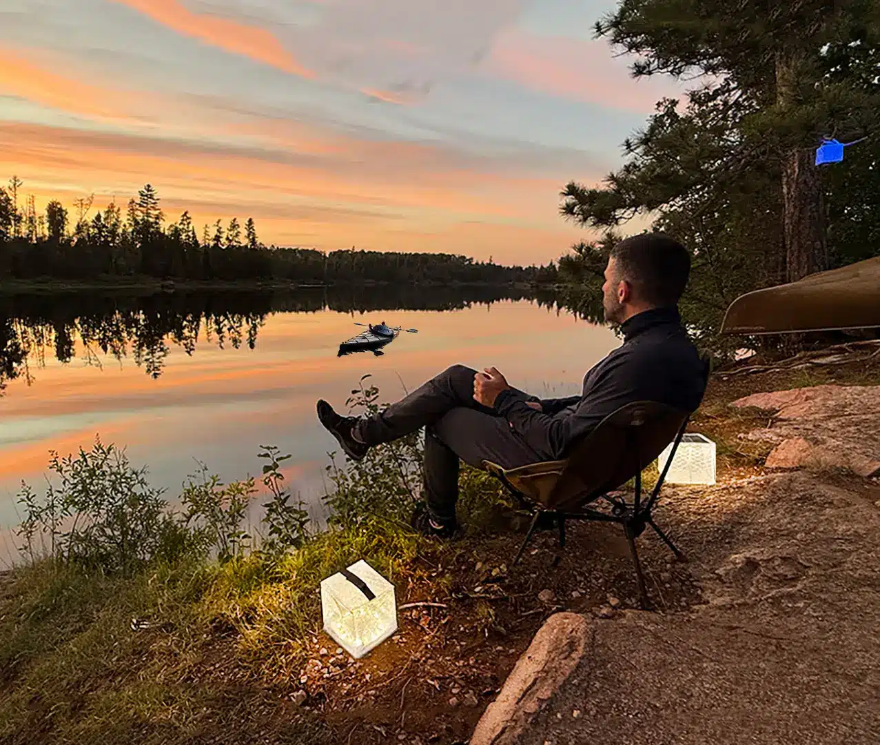 Person sitting by a lake at sunset with glowing lanterns and canoe in the background