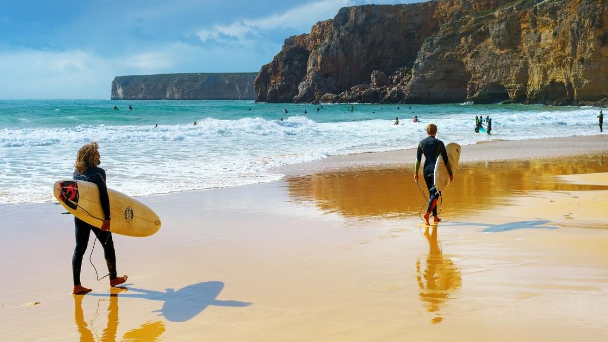 Surfers carrying boards along a sandy Portugal beach with Atlantic waves and rugged cliffs in the background.